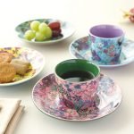 展示Nathalie Lete 陶瓷杯子 - 綠色叢林款和紫色貓款,擺放在桌子上,配有水果和餅乾,營造茶點時間的氛圍. An image of Nathalie Lete ceramic cups - Green Jungle and Purple Cat on a table with fruits and biscuits, creating a tea time setting.