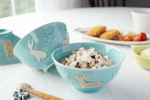 Yoshizawa 益子燒森林動物飯碗內裝米飯,並展示兩個不同側面的碗,擺放在餐桌上。 / Yoshizawa Mashiko-style forest animal rice bowl filled with rice, featuring two bowls shown from different sides on a dining table.